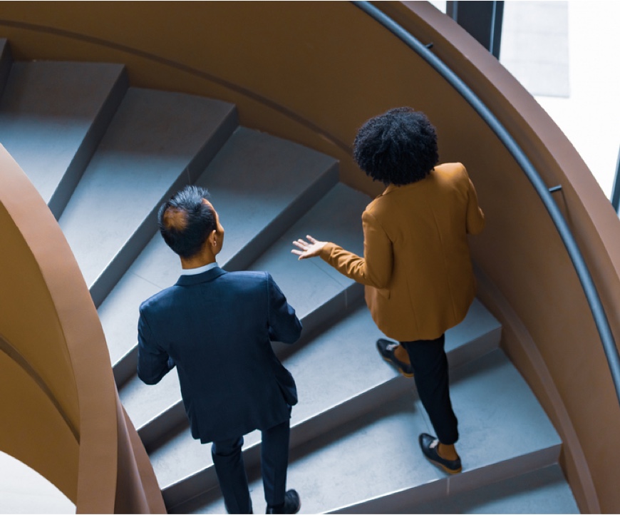 Two business professionals, a man in a suit and a woman in a blazer, walk up a spiral staircase.